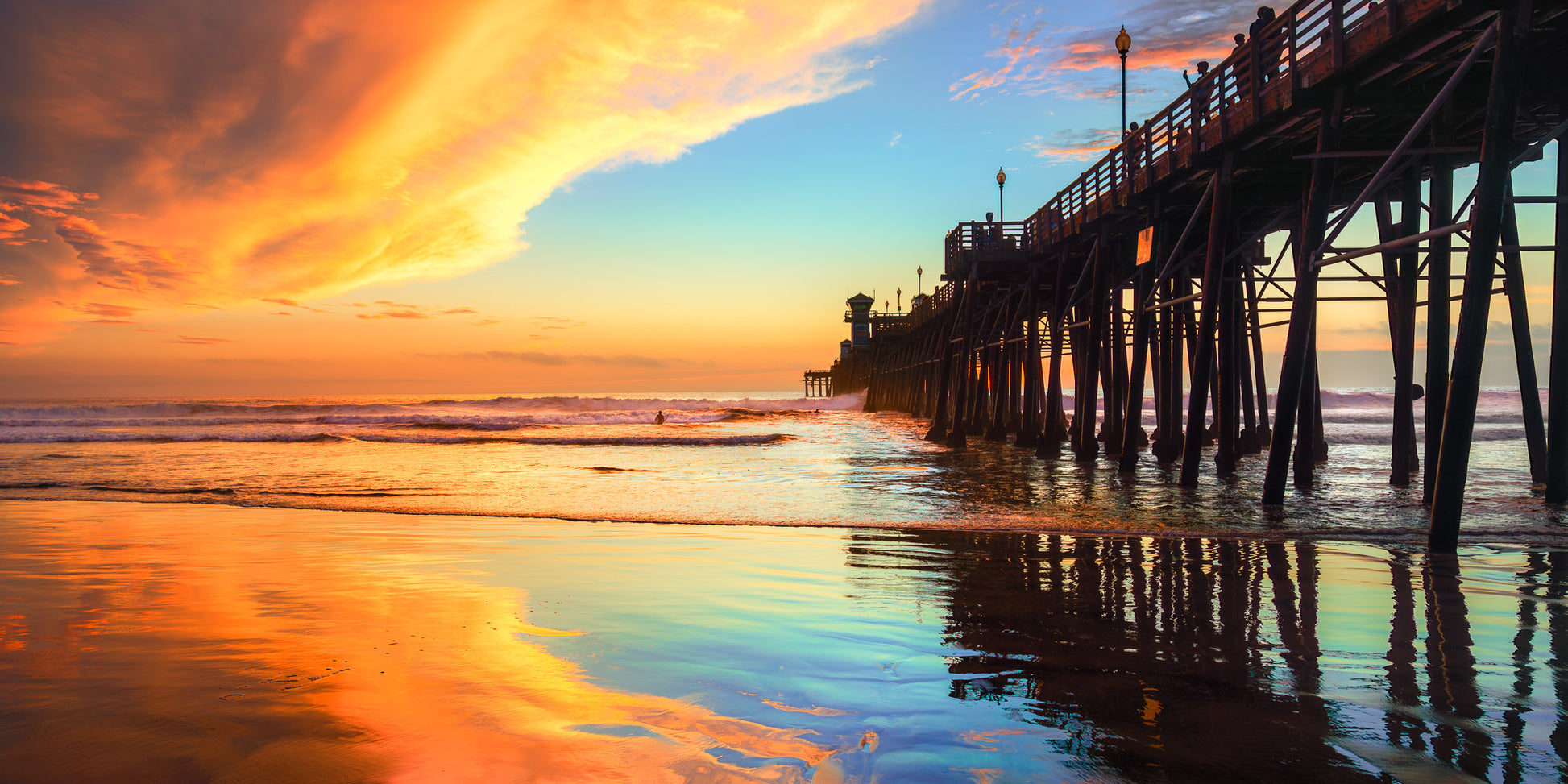 A scenic sunset over an ocean pier with dramatic orange and yellow clouds, blue sky, reflective water, and the silhouette of the pier extending into the water. "Fire n Ice" Oceanside Pier fine art photography by Craig Meyer showing dramatic contrasting lights on the pier in Oceanside California