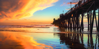A scenic sunset over an ocean pier with dramatic orange and yellow clouds, blue sky, reflective water, and the silhouette of the pier extending into the water. "Fire n Ice" Oceanside Pier fine art photography by Craig Meyer showing dramatic contrasting lights on the pier in Oceanside California