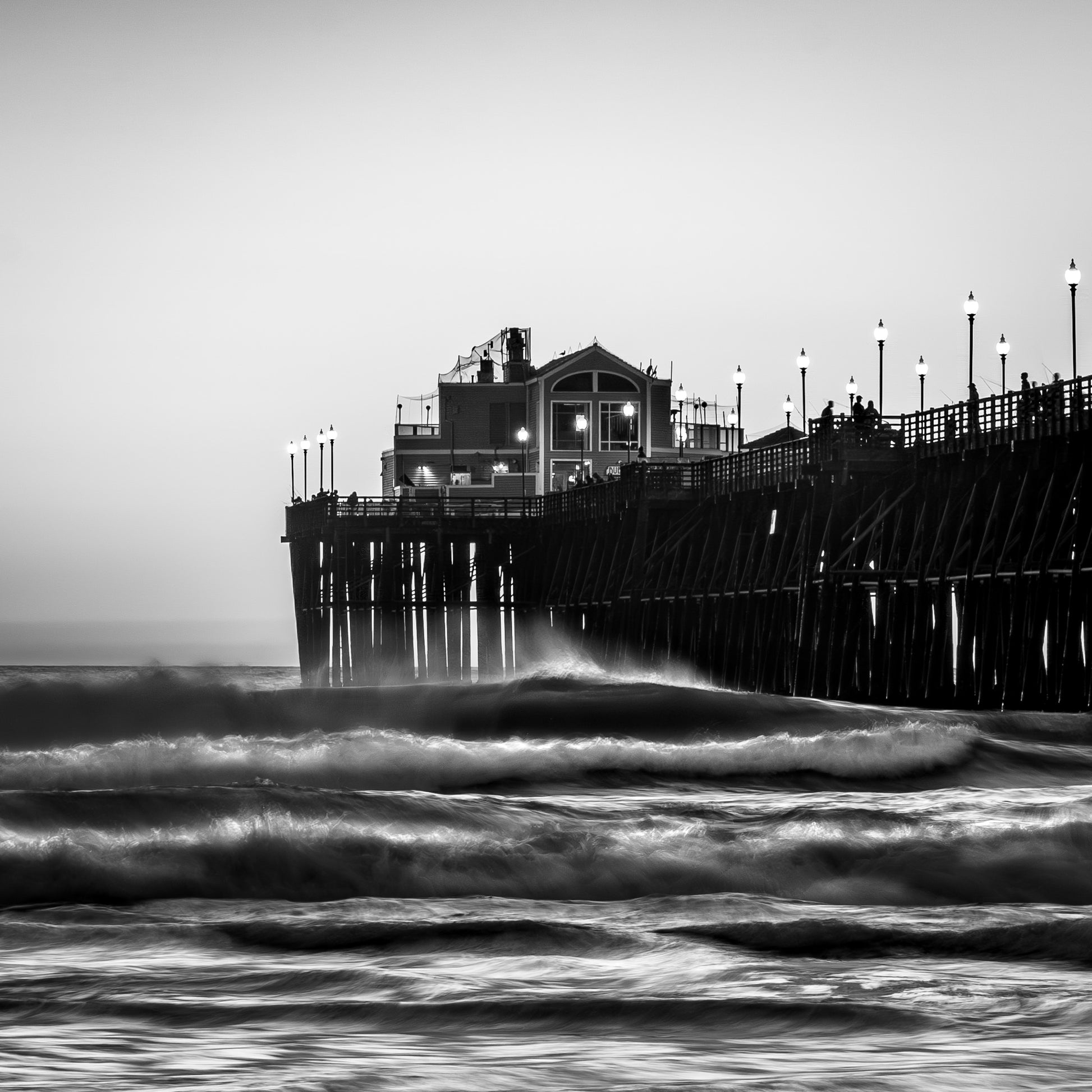 "Moonlighting" Oceanside California fine art photography by Craig Meyer showing moonlight coastal scene black and white photograph of the Oceanside pier extending into the ocean, with waves in the foreground and small figures visible on the walkway under a white sky.
